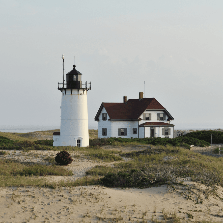 A white lighthouse beside a two-story house on a sandy landscape near the coast.