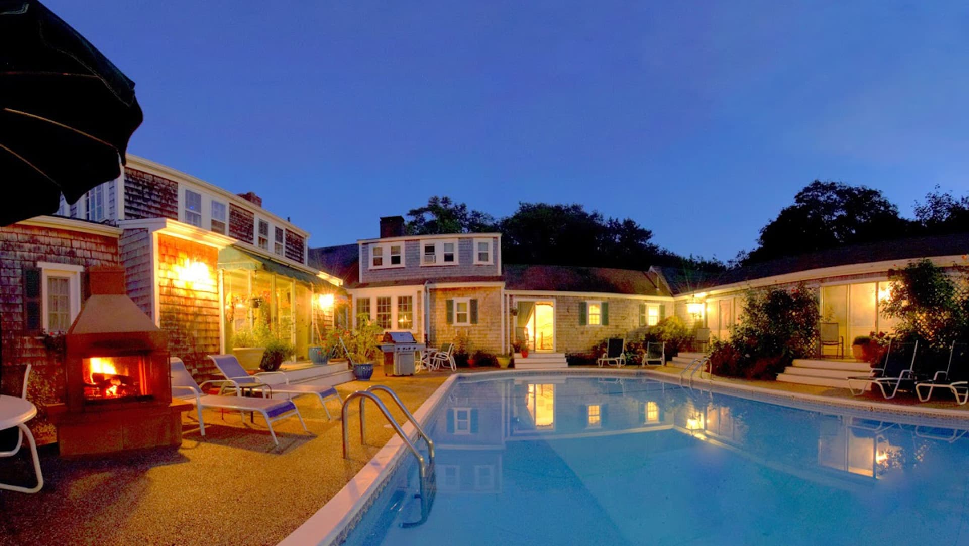 A tranquil evening scene featuring a well-lit swimming pool surrounded by cozy seating and charming wooden buildings.