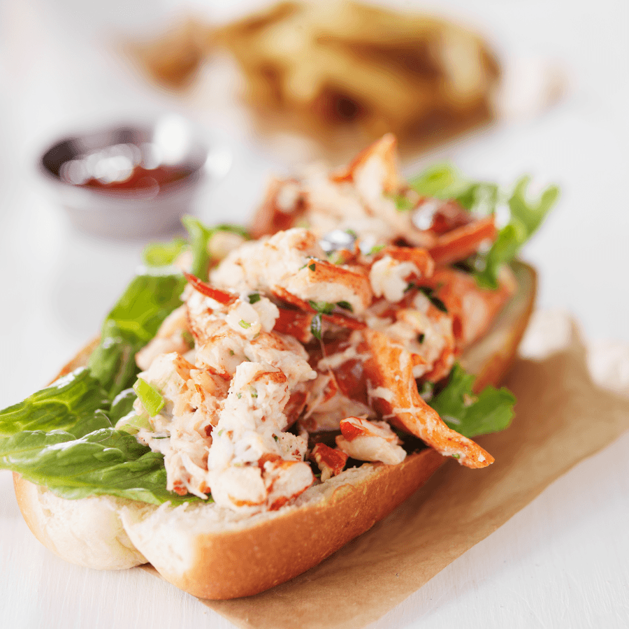 A lobster roll topped with lettuce and herbs, served alongside a small bowl of ketchup.