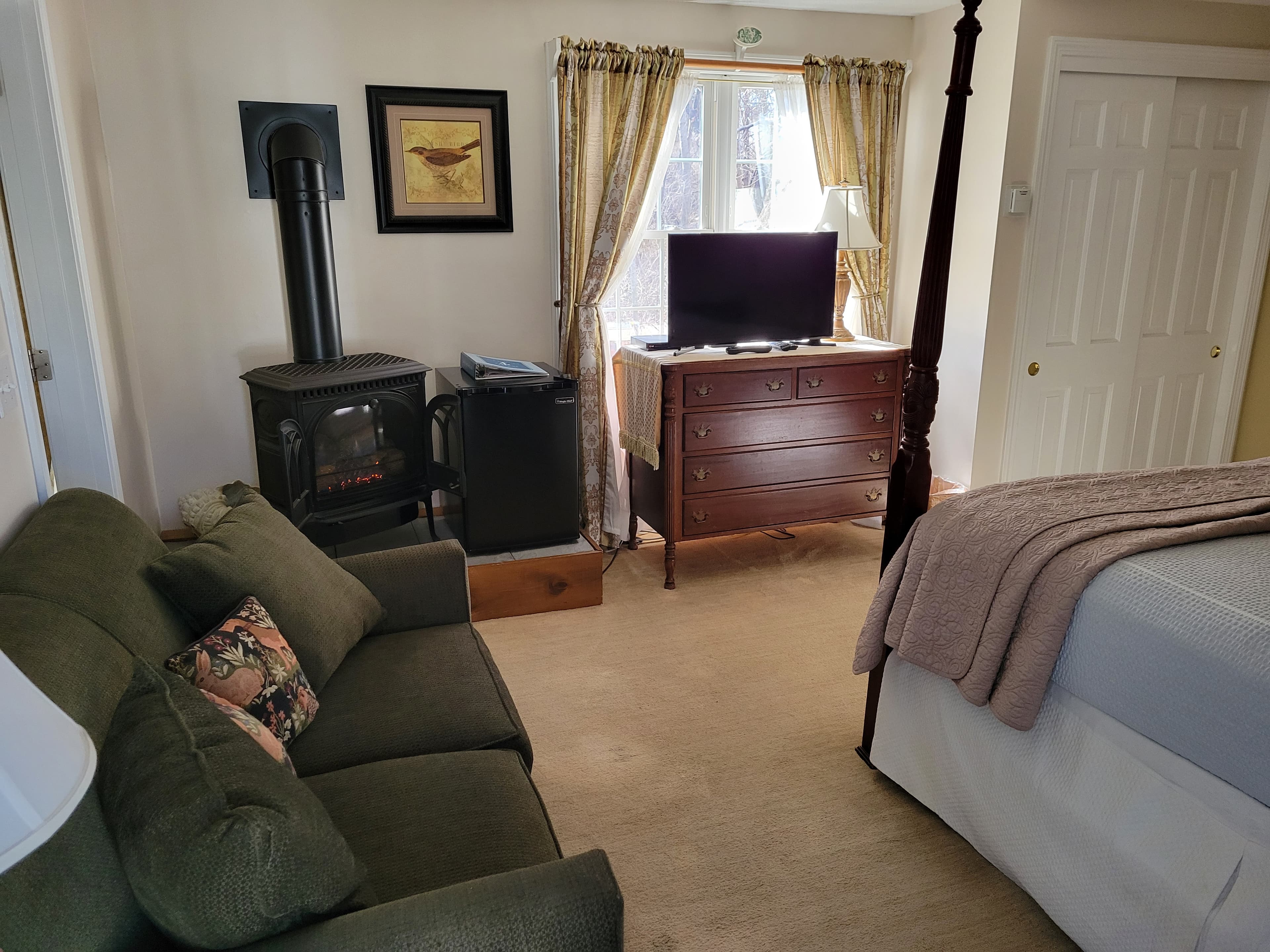Cozy bedroom featuring a green couch, a wooden dresser, a television, and a wood stove by the window.