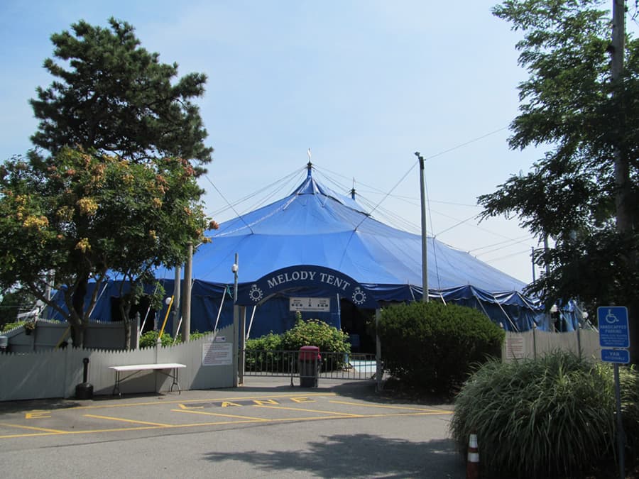 A large blue circus tent labeled "Melody Tent" surrounded by greenery and parking spaces.