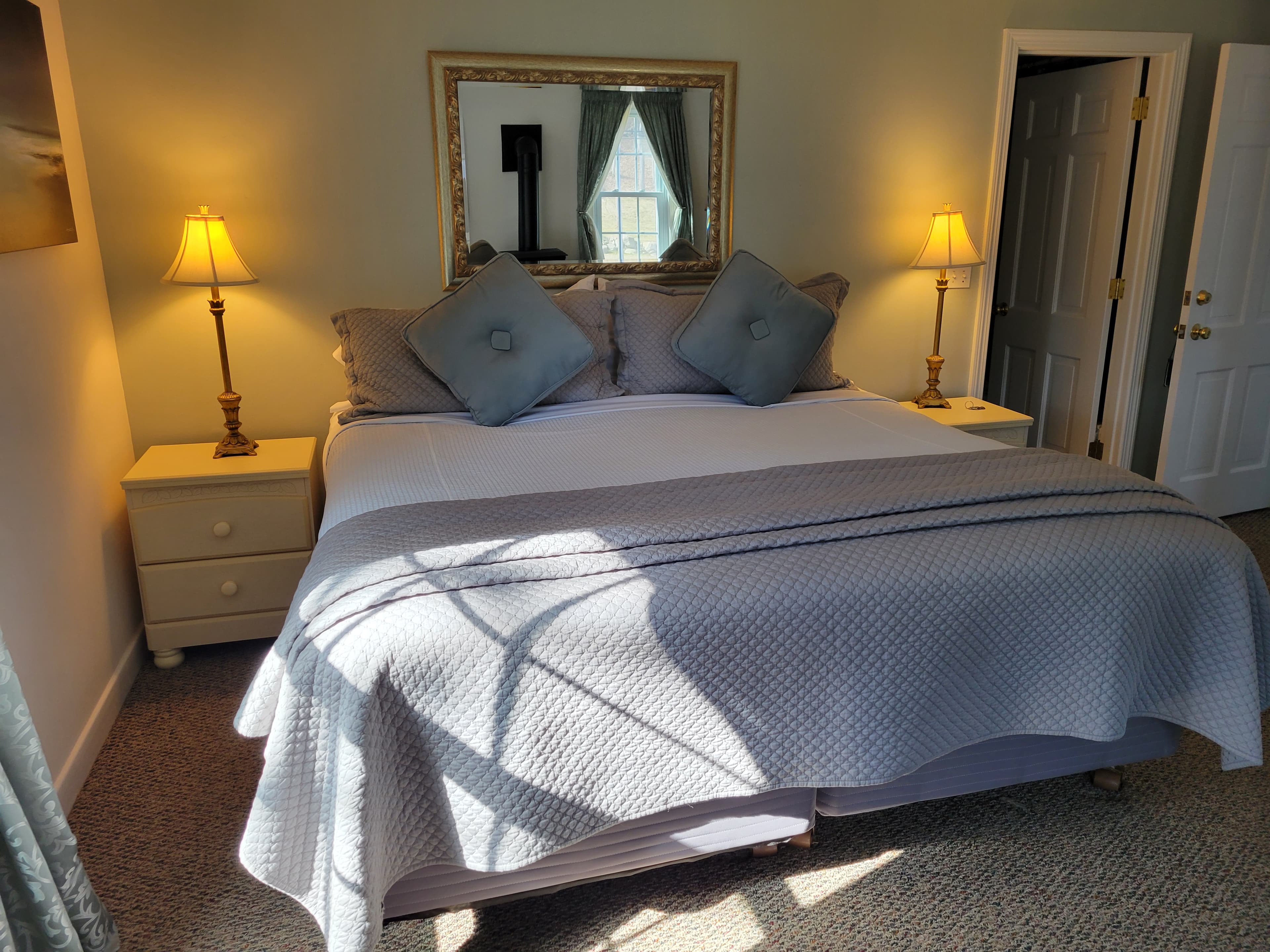 A cozy bedroom featuring a queen-sized bed with blue pillows, two bedside lamps, and a mirror reflecting natural light.