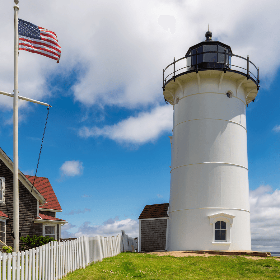 A white lighthouse stands beside a small house and an American flag under a bright blue sky.